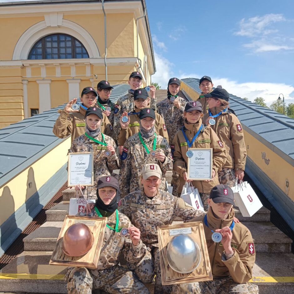 Attēlā redzami jaunieši, jaunsardzes formās ar diplomiem rokās. Pa vidu stāv instruktore Gunita Reināne