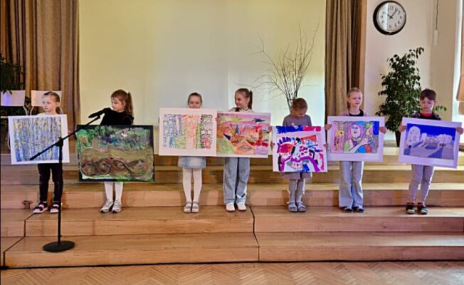 Attēlā redzami septiņi 1.klašu skolēni, kuri rāda savus uzzīmētos darbiņus uz A3 lapām