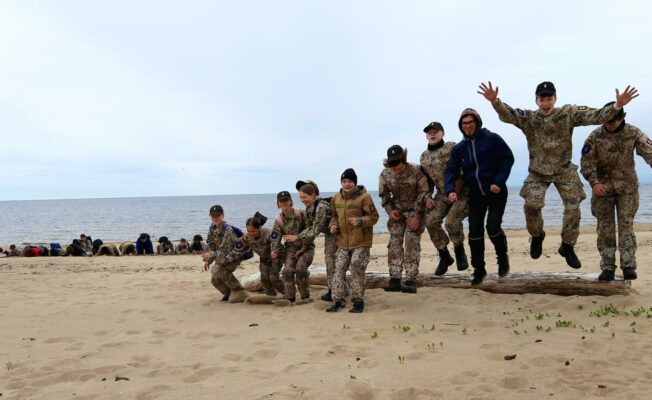 Attēlā redzami jaunsargi formas tērpos jūras malā., lecot lejā no uz zemes guļoša koka