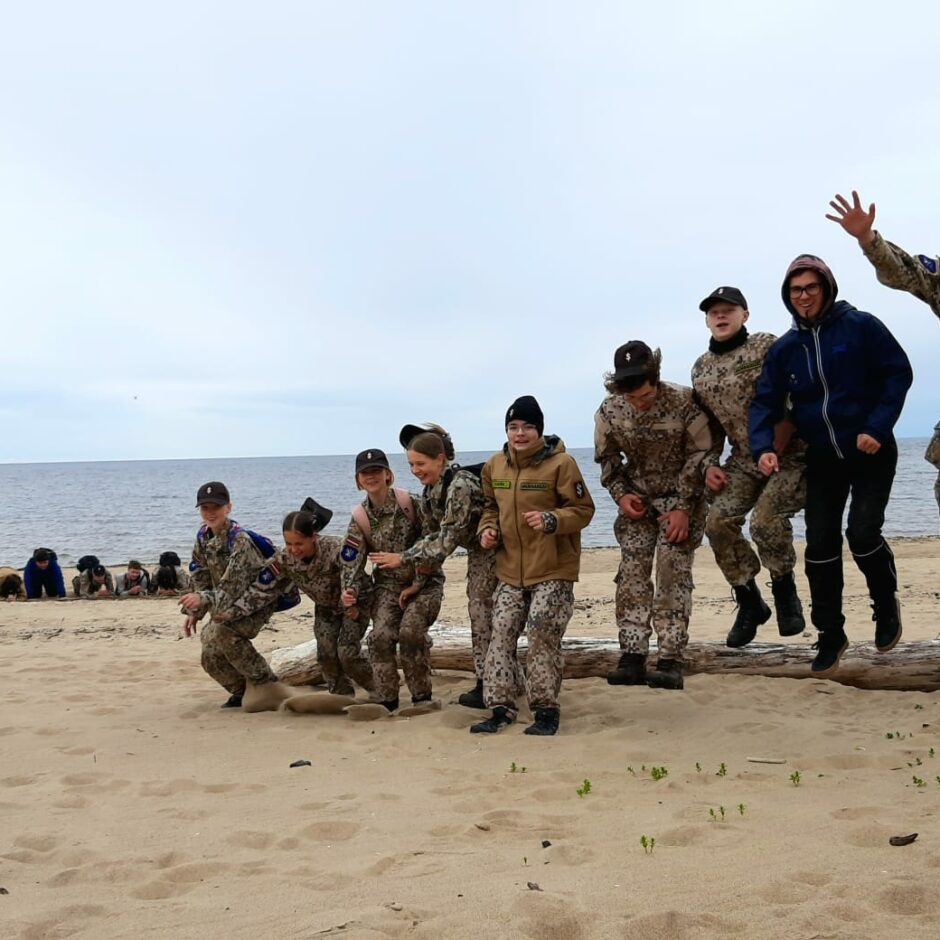 Attēlā redzami jaunsargi formas tērpos jūras malā., lecot lejā no uz zemes guļoša koka