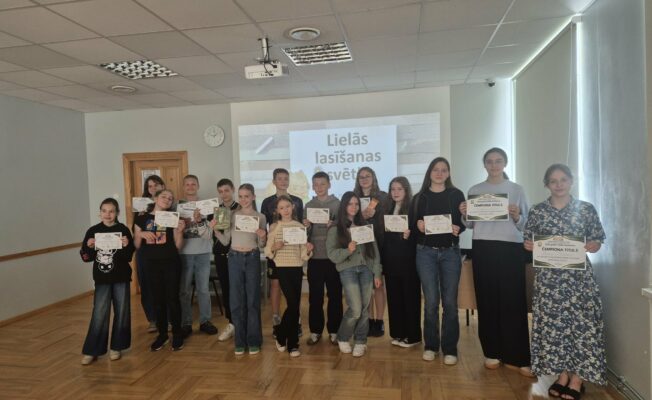 Attēlā redzami 16 skolēni, kuri stāv ar diplomiem rokās skolas mazajā zālē