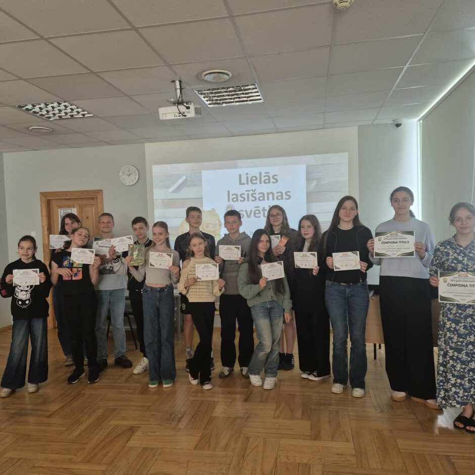 Attēlā redzami 16 skolēni, kuri stāv ar diplomiem rokās skolas mazajā zālē