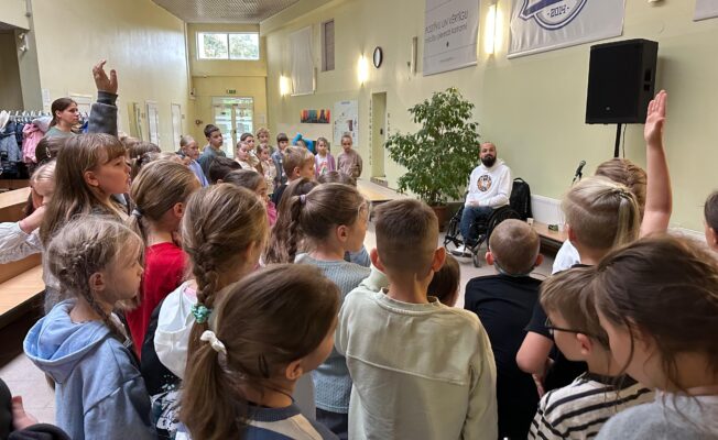 Skolēni klausās Aleksandra Lucenko stāstījumu sporta halles foajē