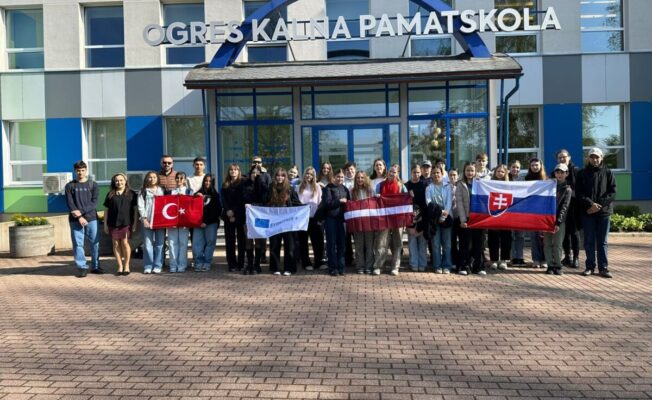 Erasmus+ mobilitātes dalībnieku kopbilde pie Ogres Kalna pamatskolas ar Turcijas, Slovākijas un Latvijas karogiem.