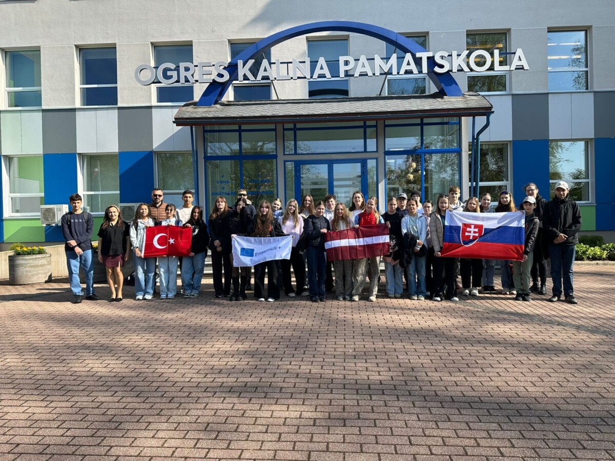 Erasmus+ mobilitātes dalībnieku kopbilde pie Ogres Kalna pamatskolas ar Turcijas, Slovākijas un Latvijas karogiem.